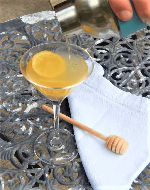 Der Bee's Knees Cocktail wird in ein Cocktailglas mit einer Zitronenscheibe gegossen. Das Glas steht auf einem Tisch mit einem hölzernen Honigstab und einer weißen Baumwollserviette.